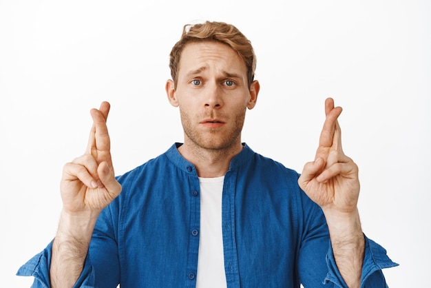 Hopeful redhead man cross fingers for good luck looking nervous and sad making wish praying for dream come true awaiting news standing worried against white background