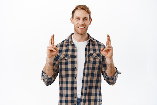 Hopeful redhead man cross fingers for good luck waiting for sign making wish anticipating positive news smiling optimistic standing over white background