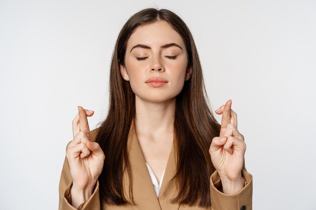 Hopeful saleswoman, corporate woman cross fingers and praying, yearning to achieve smth, standing in suit over white background. Copy space