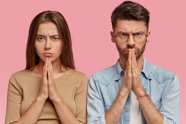 Hopeful woman and man with serious expressions keep hands in praying gesture, have gloomy look, believe in good fortune, ask God for help, isolated over pink wall. Body language concept