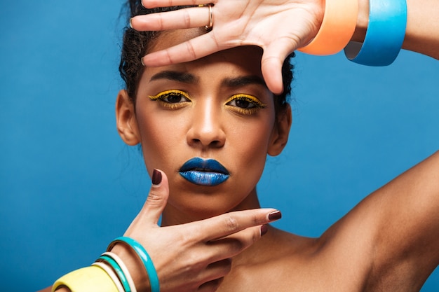 Horizontal fancy mulatto woman with colorful makeup and curly hair in bun gesturing on camera with fashion look isolated, over blue wall