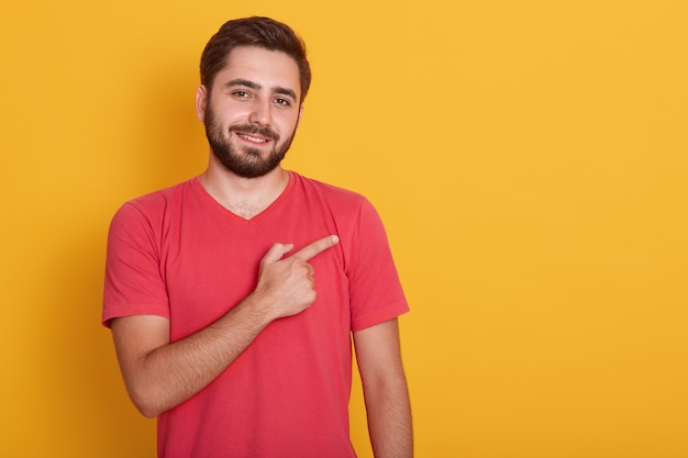 Horizontal handsome unshaven male, dressed in casual red t shirt,, pointing with index finger aside, shows copy space for your advertisment or promotion text.