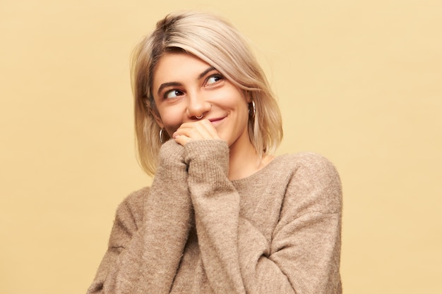 Free Photo horizontal image of attractive charming young caucasian female wearing nose ring, round earrings and stylish warm beige sweater holding hands together and looking away with mysterious smile