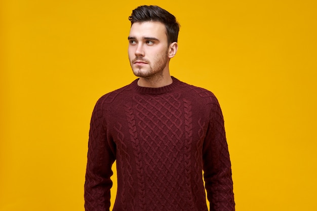 Horizontal image of attractive dark haired unshaven young Caucasian man dressed in knitted warm jumper feeling cold on winter day, looking sideways, posing isolated. People and lifestyle concept