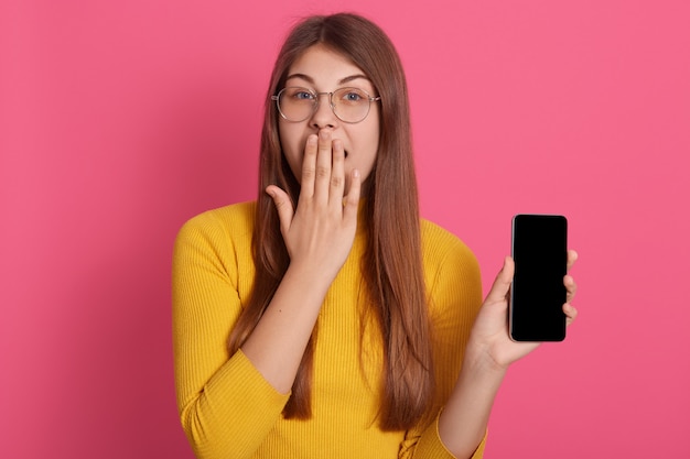 Horizontal image of beautiful young lady standing isolated over pink wall in studio, covering mouth with hand, holding smartphone, wearing eyeglasses and yellow sweatshirt. Emotions concept.