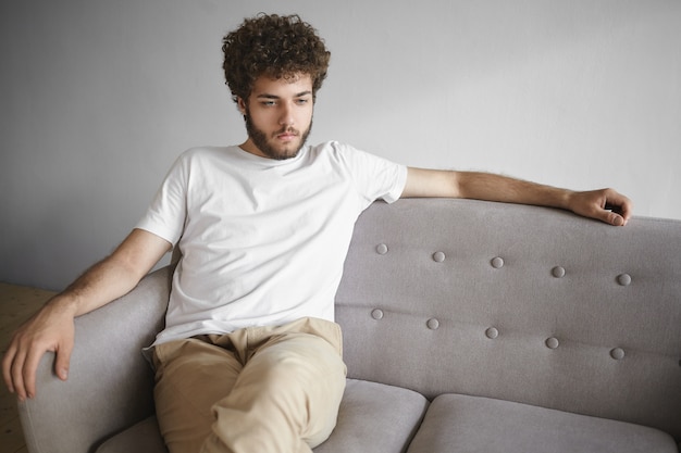 Free photo horizontal isolated portrait of attractive young caucasian man with voluminous hair and thick beard sitting casually on gray sofa at home, looking ahead of him, having pensive thoughtful expression