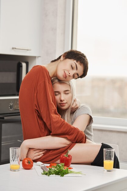 Horizontal photo of two women hugging in kitchen with closed eyes and sensual smile, sitting on table. Lovely girls in love finally opened up about their feelings to each other.