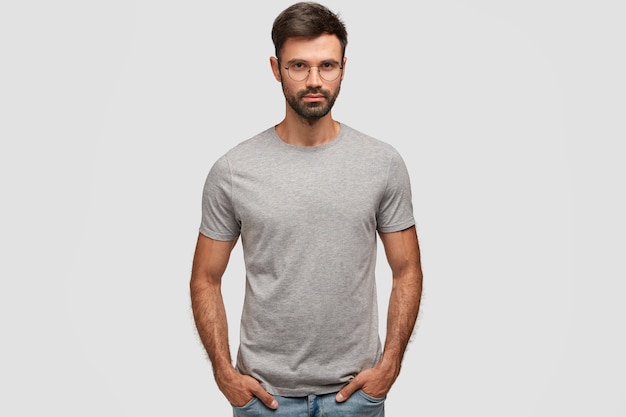 Free photo horizontal portrait of attractive bearded male with serious expression, dressed in casual grey t-shirt, keeps hands in pockets, shows new clothes, isolated over white wall. people, style concept