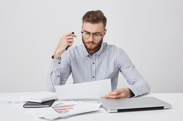 Free photo horizontal portrait of attractive beatded male manager, sits at office, surrounded with laptop