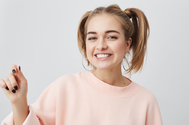 Free photo horizontal portrait of cheerful young woman with appealing smile, demonstrarting white perfect teeth, having two ponytails on her head, wearing pink sweatshirt. cute girl showing positive emotions