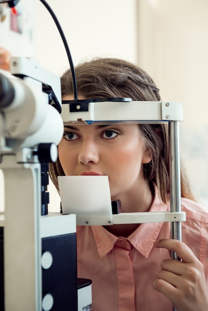 Free Photo horizontal portrait of focused european woman testing sight while looking through microbioscope, sitting in specialist office, wanting to pick appropriate glasses to see better