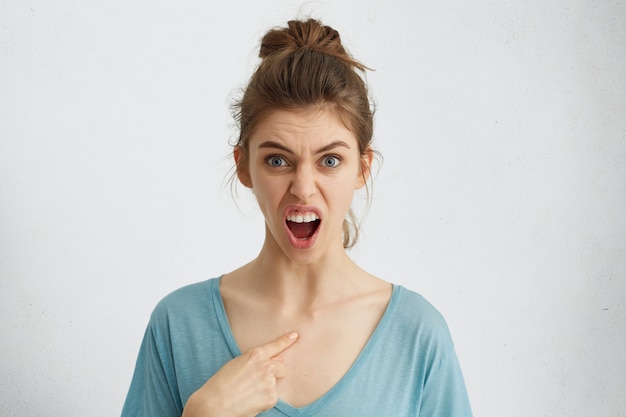 Free photo horizontal portrait of furious young woman with blue eyes having wide opened mouth from anger pointing at herself with finger frowning her eyebrows. have i done it? people and emotions concept