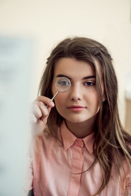 Horizontal portrait of good-looking focused woman on appointment with ophthalmologist holding lense and looking through it while trying to read word chart to check vision. Eyecare and health concept