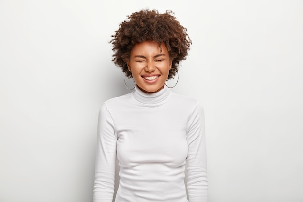 Free Photo horizontal portrait of joyous afro female laughs loudly, keeps eyes closed, being in good mood, wears turtleneck, isolated on white wall. people, satisfaction, pleasure, ethnicity, emotions