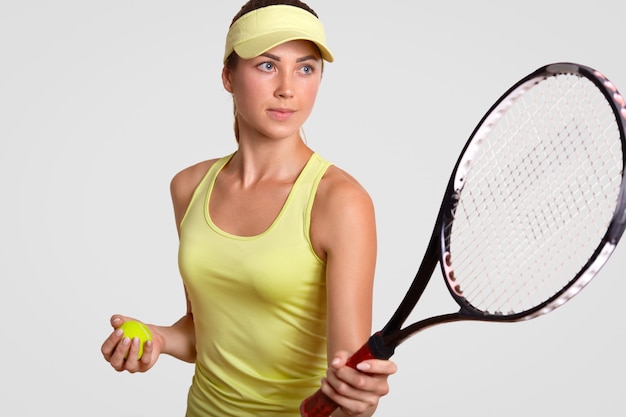 Horizontal portrait of pretty professional female tennis player holds racquet, ready to make favourite shot, holds ball