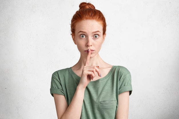 Horizontal portrait of shocked freckled female with ginger hair knot, shows hush sign, asks to keep information confidential or private, poses against white