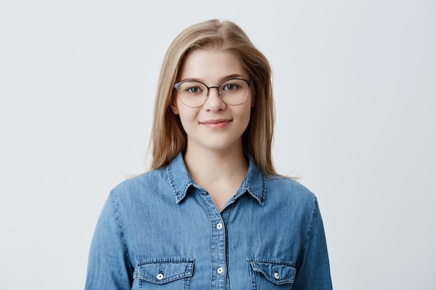 Horizontal portrait of smiling happy young pleasant looking female wears denim shirt and stylish glasses, with straight blonde hair, expresses positiveness, poses 