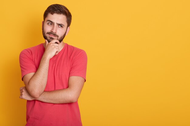 Horizontal serious unshaven male dresses casual red t shirt, keeps hand under chin, looks aside with serious facial expression, thinks about something, poses on yellow wall with free space.