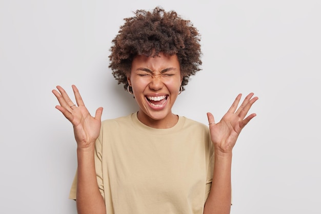 Horizontal shot of amazed happy curly woman keeps palms raised reacts to awesome news giggles positively dressed in casual brown t shirt isolated over white background Human reactions concept