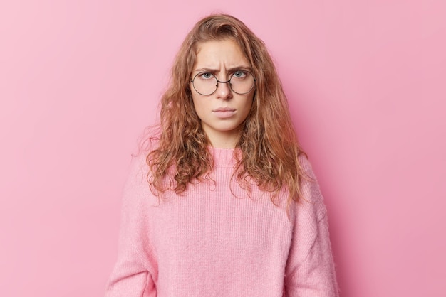 Horizontal shot of angry disappointed European woman looks unhappily at camera has grumpy expression hears something unpleasant wears round spectacles and jumper isolated over pink backgroud