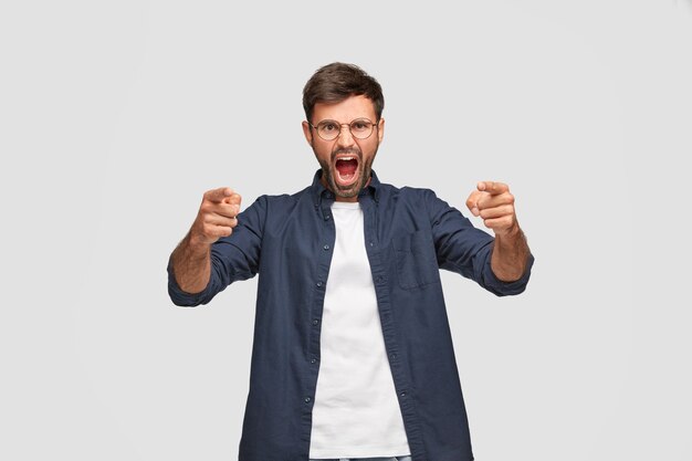 Horizontal shot of angry furious man with annoyed expression, yells at someone, points with both index fingers at camera