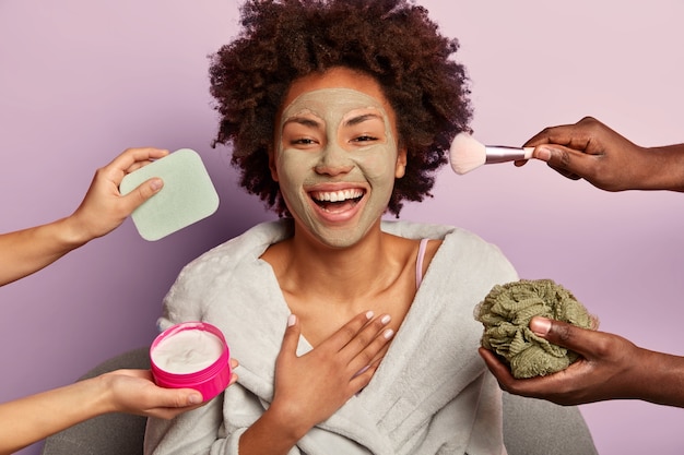 Horizontal shot of attractive female model laughs sincerely, keeps hand on chest, applies facial clay mask for skin rejuvenation, gets beauty treatments