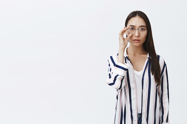 Horizontal shot of attractive natural asian woman with tanned skin and long dark hair, touching round glasses on eyes, wearing stylish striped shirt over white t-shirt, gazing dreamy