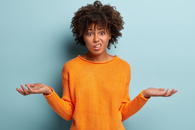 Horizontal shot of attractive woman with puzzled expression, spreads hands with confused look, frowns face, wears orange clothes, feels hesitant, indignant isolated over blue wall. Doubt concept