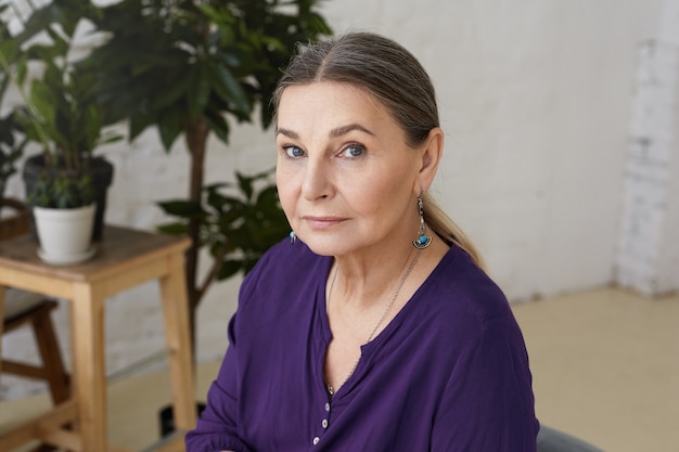 Free Photo horizontal shot of beautiful blue eyed grandmother of european appearance relaxing at home, waiting for her grandchildren, wearing violet casual shirt and earrings, having serious expression