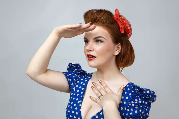 Horizontal shot of beautiful glamourous pin up girl wearing bright make up and retro dress with low cut holding palm at forehead and looking into distance trying to see something