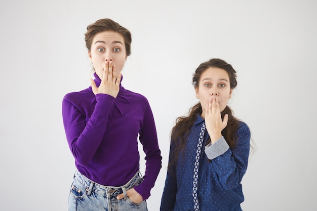 Free photo horizontal shot of beautiful stylish 20 year old woman and her teenage sister expressing astonishment and full disbelief, covering mouths