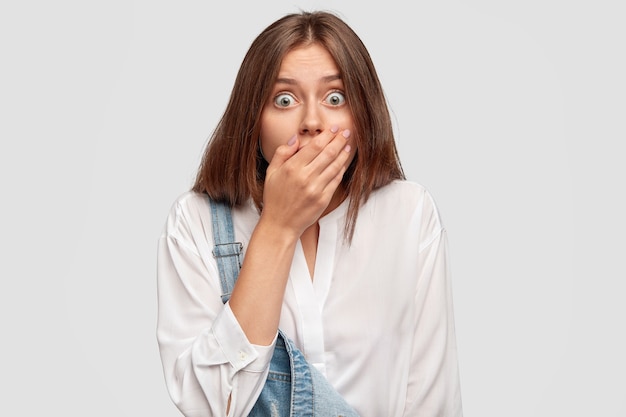 Free photo horizontal shot of bug eyed terrific european female covers mouth, recieves awful news, dressed in fashionable white shirt and denim overalls, isolated over wall. people, amazement concept