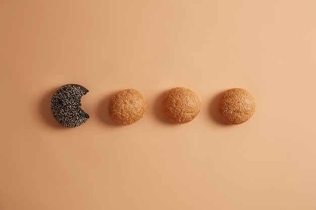 Horizontal shot of burger buns in row, one black cuttlefish ink is bitten, ready for making burgers or sandwiches. Unhealthy food and junk nutrition concept. Copy space for your information.