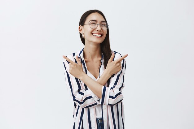 Horizontal shot of carefree sociable and friendly stylish woman in glasses and striped blouse, crossing hands and pointing at different sides, looking right with joyful happy smile