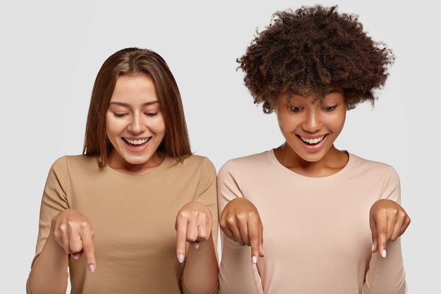 Free photo horizontal shot of cheerful mixed race two young women point at floor with fore fingers, dressed in casual outfit