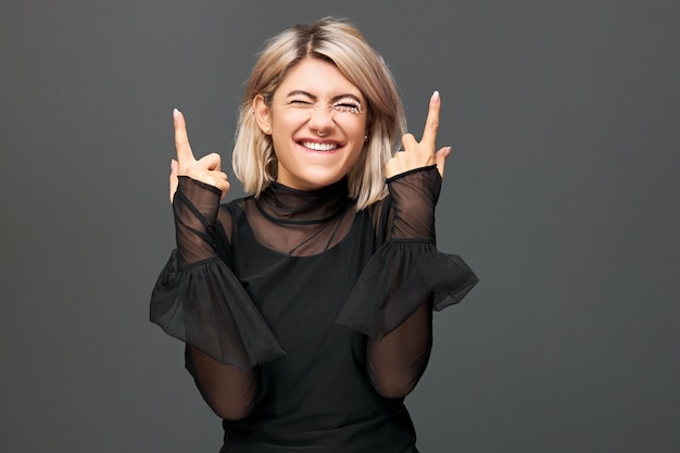 Horizontal shot of cheerful overjoyed ecstatic young European woman wearing stylish transparent dress enjoying good positive news, expressing joy, pointing index fingers up, celebrating success