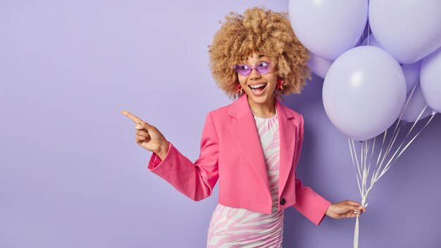 Horizontal shot of cheerful woman dressed in fashionable clothes holds bunch of inflated balloons celebrates university leaving organizes party points away on copy space isolated on purple background