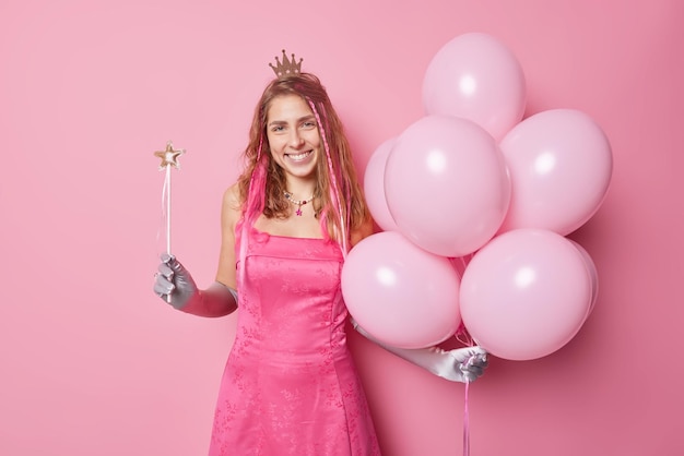 Horizontal shot of cheerful woman prepares for celebration wears festive dress long gloves crown holds magic wand and bunch of inflated balloons isolated over pink background Holidays concept