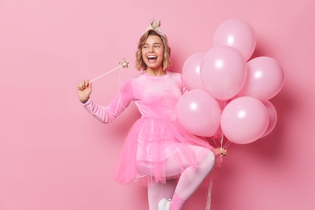 Free photo horizontal shot of cheerful young blonde woman dressed in festive outfit holds magic wand and bunch of balloons celebrates birthday glad to meet guests on party poses against pink background