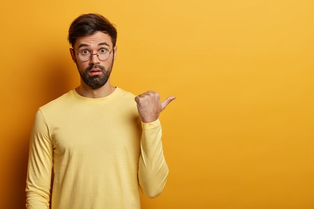 Free photo horizontal shot of confused bearded man points thumb right, discusses discount offer, tells about amazing unbelievable sale, wears glasses and sweater, copy space for your promotional content