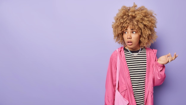 Free photo horizontal shot of confused hesitant curly woman shrugs shoulders feels uncertain wears casual striped jumper and pink jacket feels bewildered isolated over purple background blank copy space