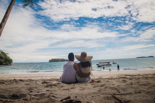 Horizontal shot of a couple sitting on a beach toward the calm blue water under the beautiful sky