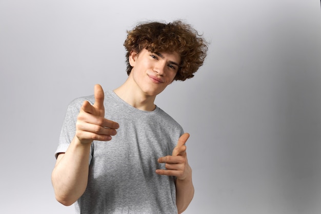 Horizontal shot of cute confident young man with smooth face and curly hairdo wearing casual clothing posing against gray background pointing fore fingers at camera, smiling, being proud of you