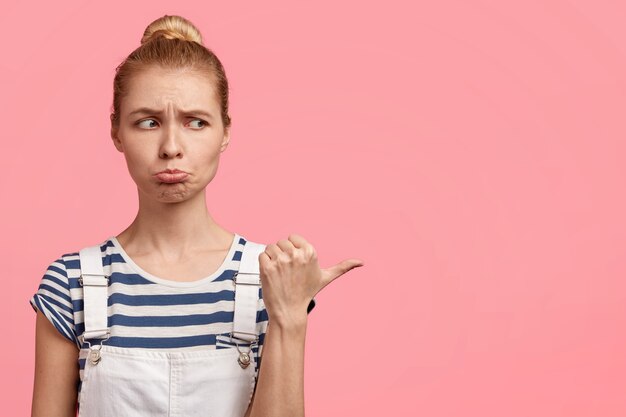 Free photo horizontal shot of displeased gloomy blonde female purses lips and points aside with thumb, feels discontent with something, poses over pink wall