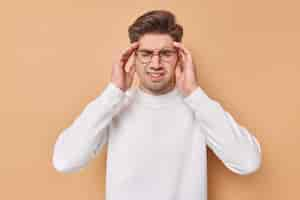 Free photo horizontal shot of displeased young man suffers from migraine keeps hands on temples has severe headache tries to reveal pain wears spectacles and white jumper isolated over beige background