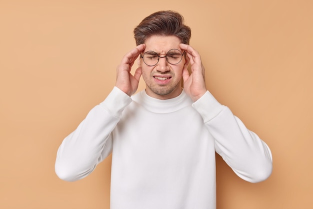 Free Photo horizontal shot of displeased young man suffers from migraine keeps hands on temples has severe headache tries to reveal pain wears spectacles and white jumper isolated over beige background