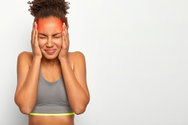 Free photo horizontal shot of dissatisfied curly haired woman touches both temples, suffers from migraine, wears sports bra, closes eyes from bad feelings, isolated on white wall