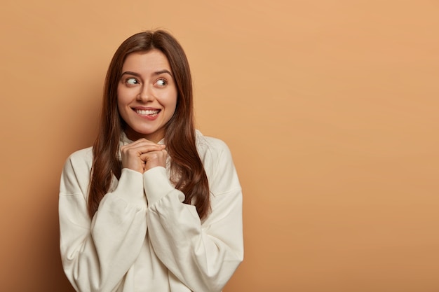 Horizontal shot of dreamy woman anticipates for something pleasant, bites lips, keeps hands together, concentrated aside, has delighted expression