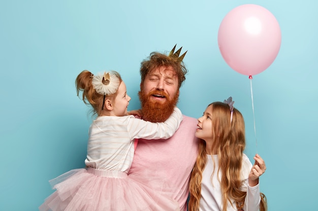 Free photo horizontal shot of fatigue young bearded man with ginger hair, tired of playing with children. two daughters spend holiday together with affectionate dad, hold pink balloon. festive day concept