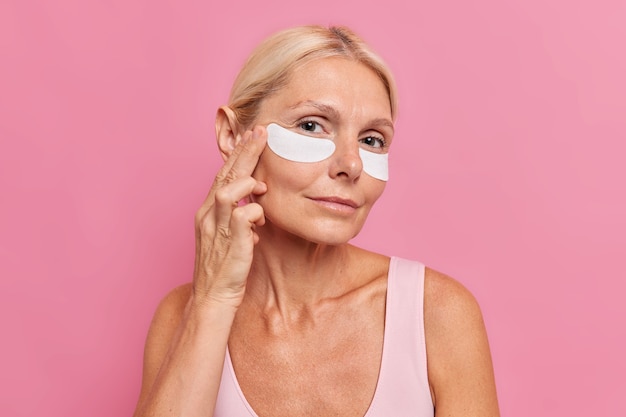 Free Photo horizontal shot of forty years old blonde woman applies beauty patches under eyes undergoes anti aging procedures looks attentively at camera isolated over pink wall